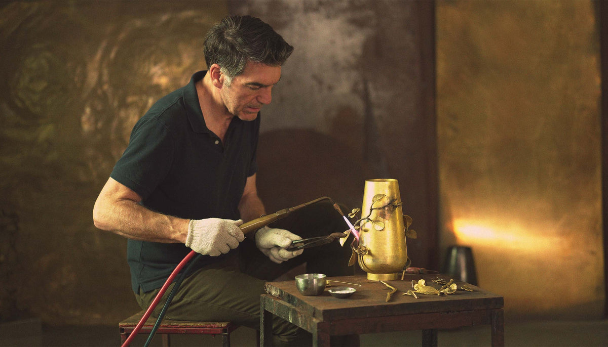 A man wearing a dark green polo shirt and white gloves sits at a small table, using a torch with a bright flame to work on a gold-colored metal vase or decorative object against a warm, textured brown background.