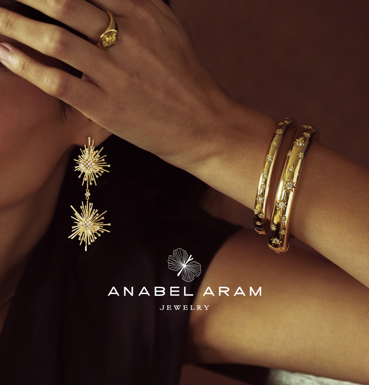 A close-up lifestyle shot of a model wearing gold jewelry from Anabel Aram Jewelry, featuring a starburst drop earring, a matching ring, and stacked bangles on her wrist, set against a dark background with the brand logo at the bottom.