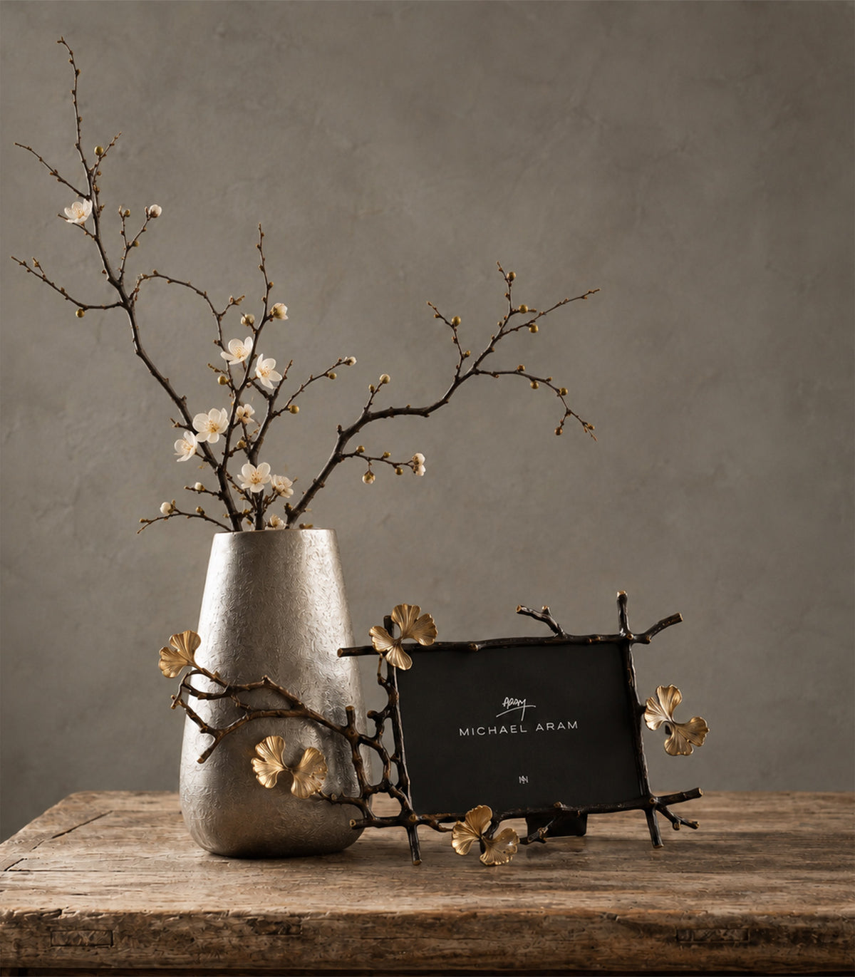 A Michael Aram decorative picture frame with gold leaf petals and branch details, styled next to a textured gray ceramic vase with white blossoms on a rustic wooden table against a textured gray wall.