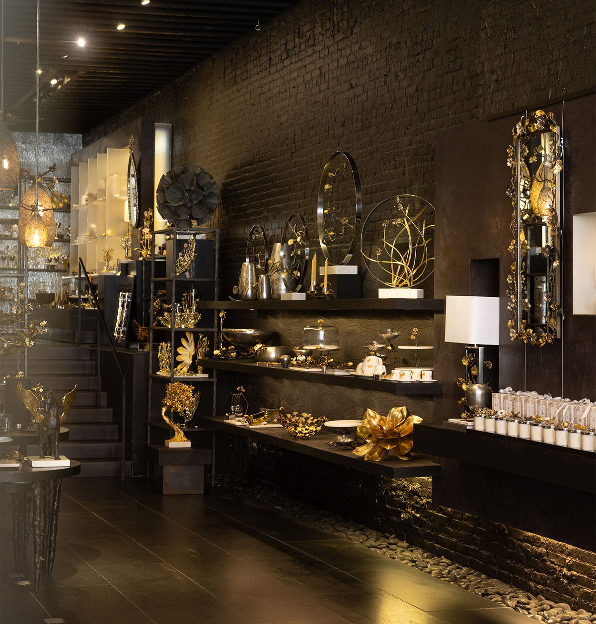 A luxury interior display of dark wood and gold decorative home goods, including ornate mirrors, gold leaf sculptures, a table lamp, and glassware, set against a dark brick wall.