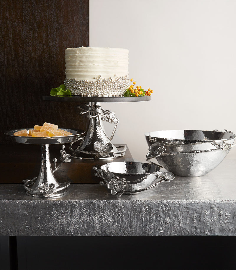A set of metallic silver serving pieces, including a tiered cake stand with a dragon motif, a pedestal plate with candy, a shallow bowl, and a larger bowl, arranged on a textured gray table against a split dark wood and white wall background.